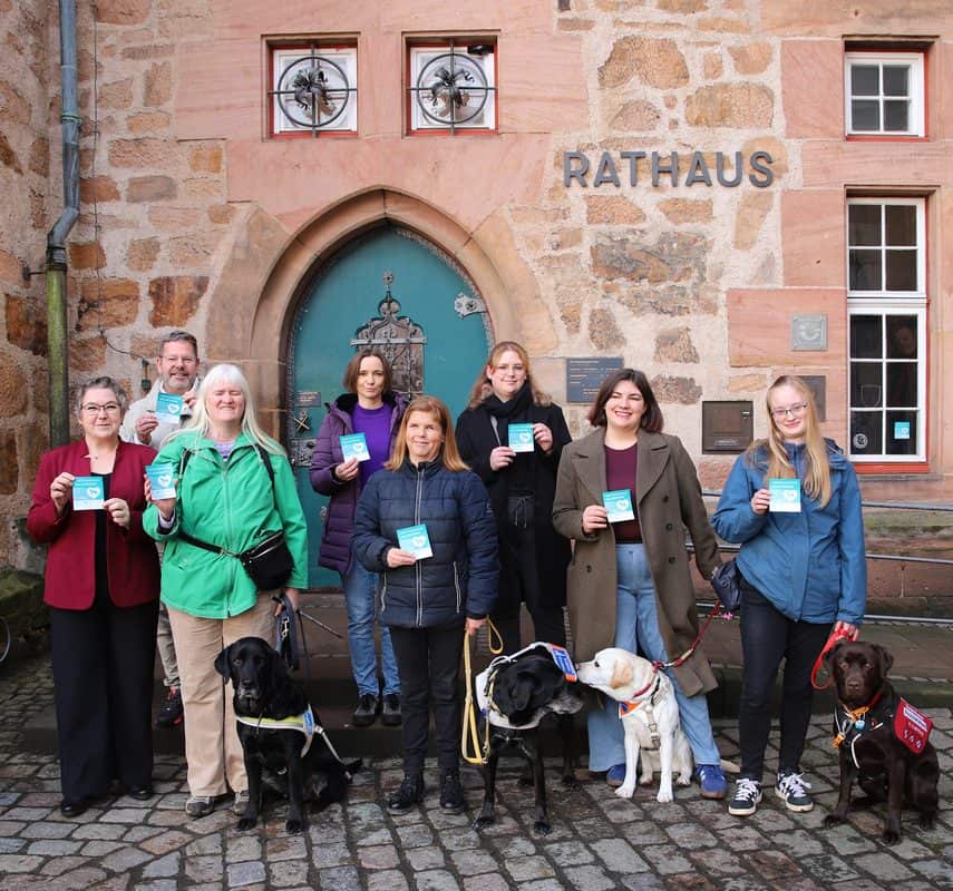Neun Menschen stehen vor dem historischen Marburger Rathaus mit seiner türkisfarbenen Spitzbogentür und halten Aufkleber mit der Aufschrift „Assistenzhund willkommen“ in die Kamera. Vor ihnen sitzen vier Hunde: zwei schwarze Labradore im Führgeschirr, ein heller Labrador und ein brauner Labrador mit roter Kenndecke „Assistenzhund in Ausbildung“.
