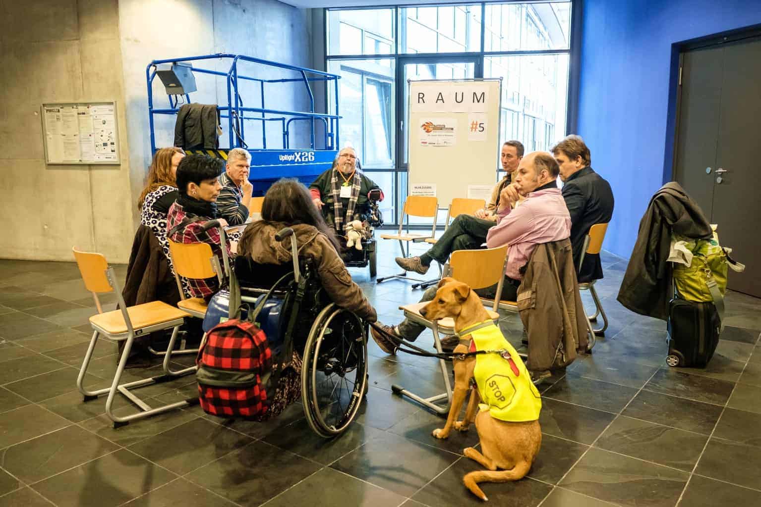 Photo of a working meeting :: A group of eight adults sits together in a circle in a room, seemingly engrossed in a work conversation. A wheelchair user is accompanied by an assistance dog sitting in the foreground of the image.