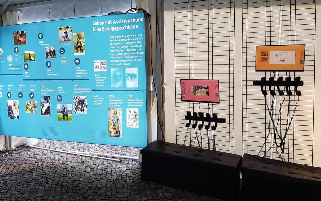 Foto Assistenzhunde Ausstellung :: Auf dem Foto sieht man ein Bild der Assistenzhunde-Ausstellung.
Auf einer Fotowand ist die