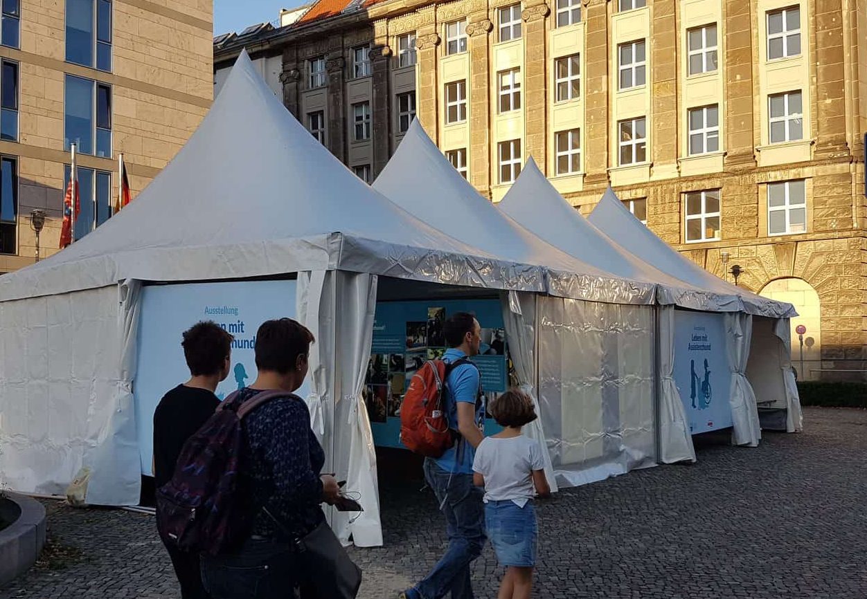 Foto der Ausstellungszelte :: Auf dem Foto sieht man vier Menschen vor den Ausstellungszelten der Assistenzhunde-Ausstellung entlang gehen.