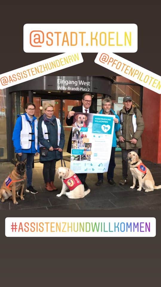 Aufkleberaktion Köln :: Gruppenbild der Aktion am Stadthaus Deutz. Stadtdirektor Dr. Keller und Behindertenbeauftragter der Stadt Köln, Dr. Bell halten das Poster hoch, das im DIN A3 Format zusammen mit den Aufklebern in den städtischen Behörden angebracht werden wird. Sie sind umrahmt von AssistenzhundhalterInnen Julia S., Manja M. und Günter M. Im Vordergrund die Assistenzhunde.
Weiterhin ist das Bild dekoriert mit #AssistenzhundWillkommen @Köln – unsere Stadt @Assistenzhunde NRW @PfotenPiloten
Bildquelle Julia S. 