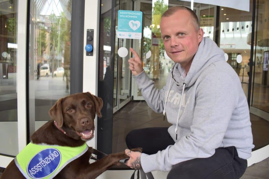 Ein Mann hockt mit seinem Assistenzhund vor einer Eingangstür, an der ein Aufkleber der Zutrittskampagne "Assistenzhund Willkommen" klebt.