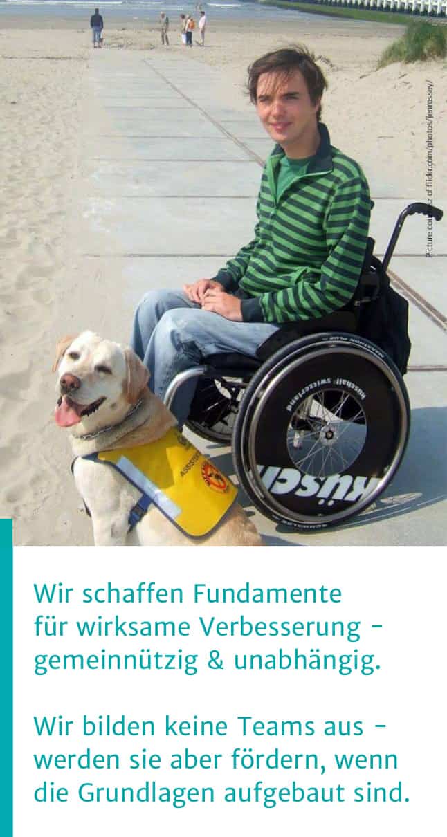 Assistenzhund TJ mit Halter am Strand :: Rechts im Bild ein junger Mann im Rollstuhl an weitem Sandstrand. Im Hintergrund das Meer und Menschen. Im Vordergrund sieht ein freundlicher, heller Labrador mit gelber Kenndecke in die Kamera. Darunter der Text: Wir schaffen Fundamentefür wirksame Verbesserung - gemeinnützig & unabhängig.Wir bilden keine Teams aus -
werden sie aber fördern, 
sobald die Grundlagen stehen.