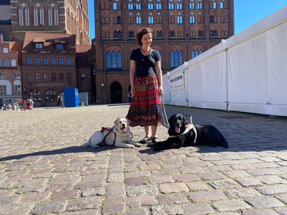 Hannah is standing in front of Stralsund City Hall with guide dog Daika and medical assistance dog Mascha at her feet.
