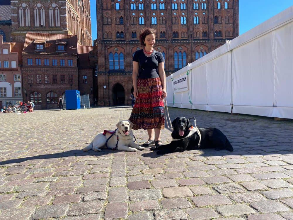 Hannah steht vor dem Stralsunder Rathaus und zu ihren Füßen liegen Führhündin Daika und medizinischer Assistenzhund Mascha.