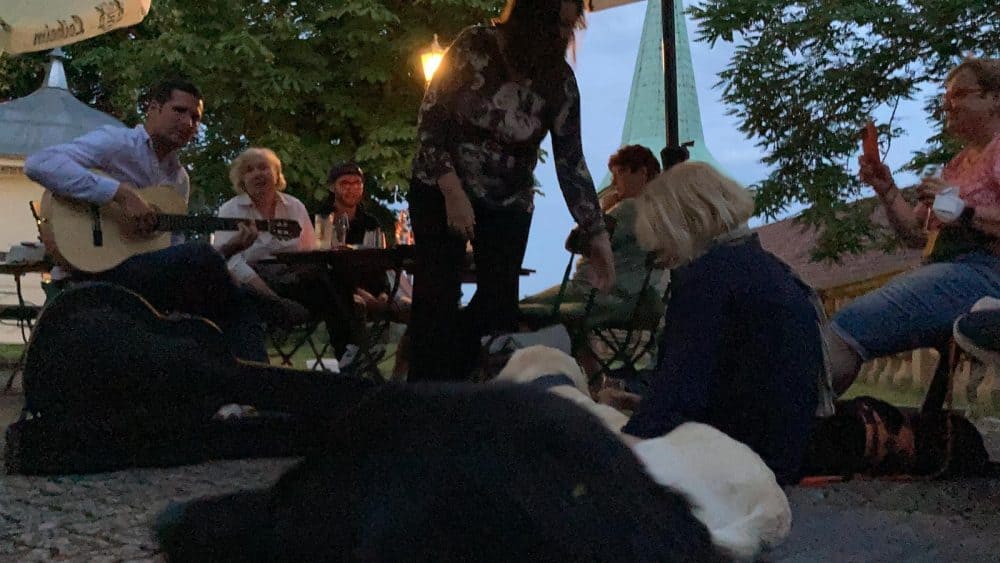 Es ist fast dunkel, Peter Levay spielt Gitarre, es sitzen Menschen bei den Hunden auf dem Boden, andere am Tisch und auf einer nahen Bank.