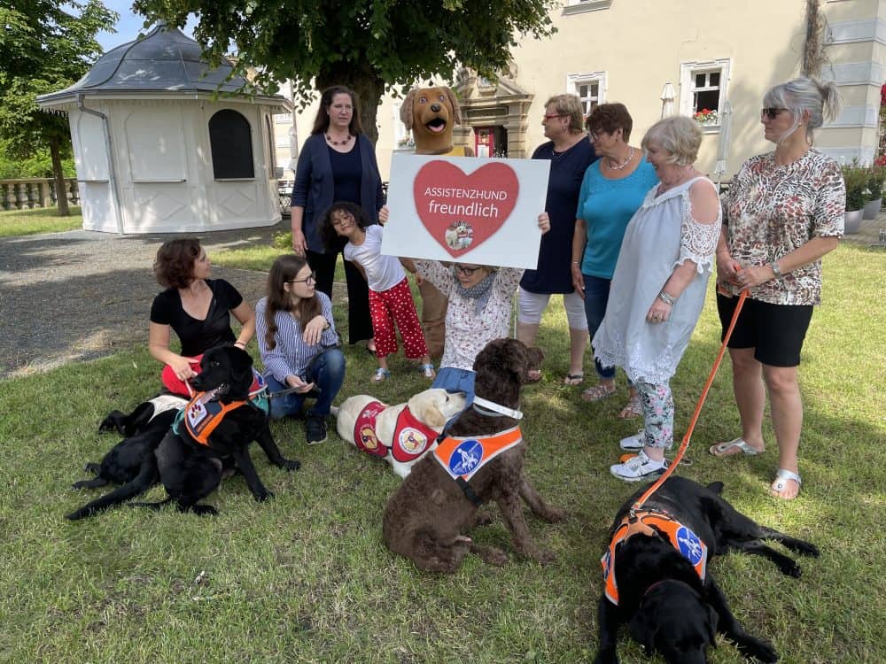 Die TeilnehmerInnen posieren mit ihren Assistenzhunden für ein Gruppenfoto mit Maskottchen Benji und einem Schild "assistenzhundfreundlich".