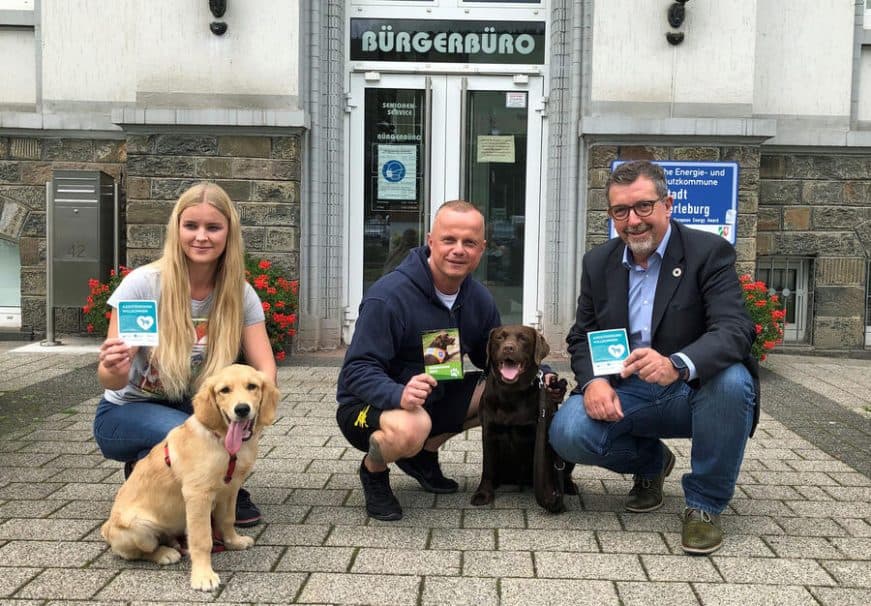 Gemeinsam mit Assistenzhündin "Hanni" brachten Thorsten Habel und Bad Berleburgs Rathausmitarbeiterin Sarah Müsse mit Bürohund "Carlo" das sichtbare Zeichen (Aufkleber) für die Zutrittskampagne "Assistenzhund Willkommen" am Eingang zum Bürgerbüro an. Alle hocken sich neben die beiden Hunden.