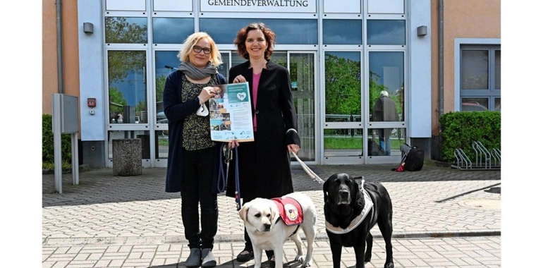 Manja Myrrhe-Kohlenbrenner mit Mascha (blonde Labradorhündin mit rote Assistenzhundveste) und Hannah Reuter mit Daika (schwarze Labradorhündin im Führgeschirr).