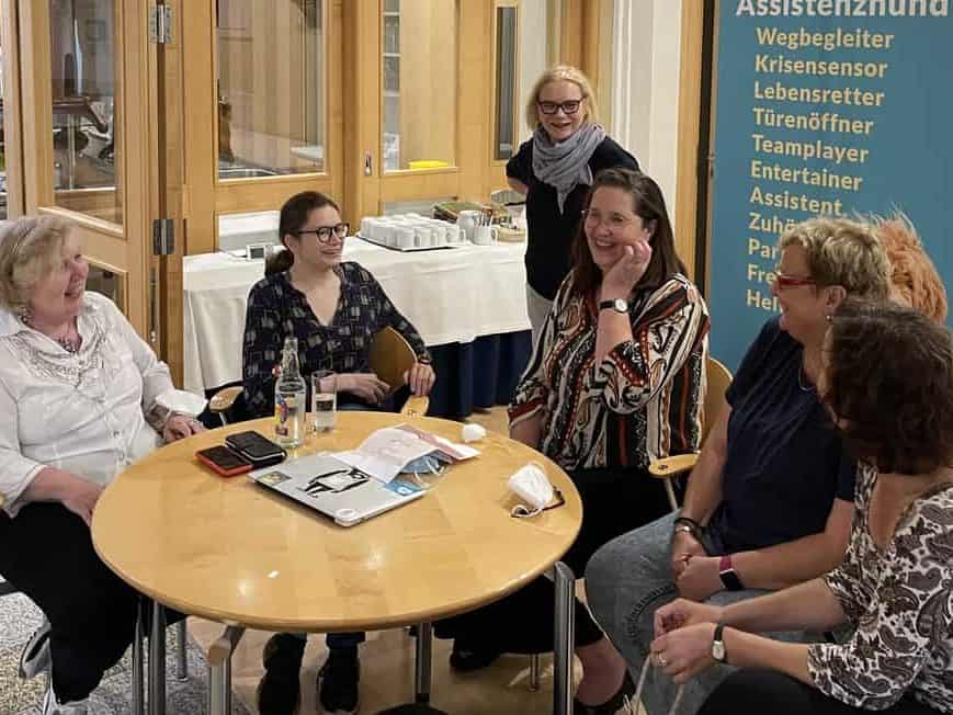 Eine Gruppe Assistenzhundhalterinnen sitzt mit R. Warda an einem runden Tisch im Seminarraum berät sich und hat offensichtlich Spaß.