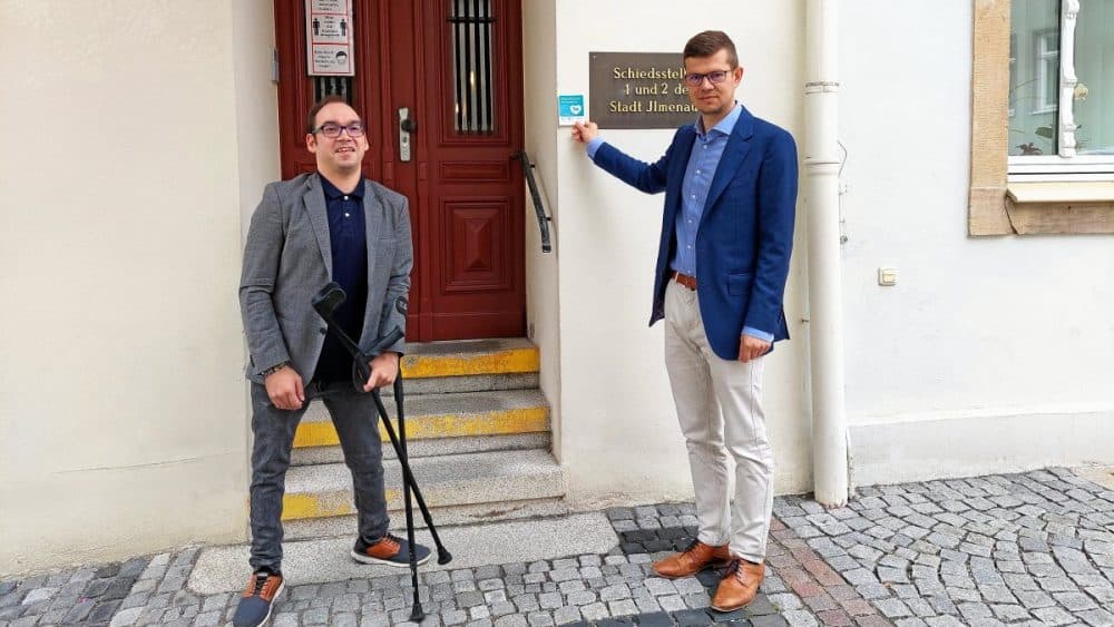 Ilmenaus Inklusionsbeauftragter Philipp Schiele und Oberbürgermeister Daniel Schultheiß (von links) setzen sich für die Kampagne „Assistenzhund Willkommen“ ein. Schultheiß präsentiert den Sticker, den an der Aktion teilnehmende Händler an die Tür kleben, um zu zeigen, dass Assistenzhunde mit ins Gebäude dürfen. Zudem liegt bei diesen Händlern Informationsmaterial aus.