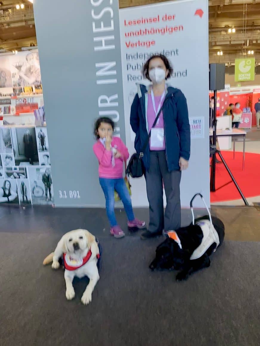 Vor dem Schild der unabhängigen Verlage stehen Hannah mit Mila und Daika. Assistenzhund Mascha hat sich mit auf das Bild geschummelt.