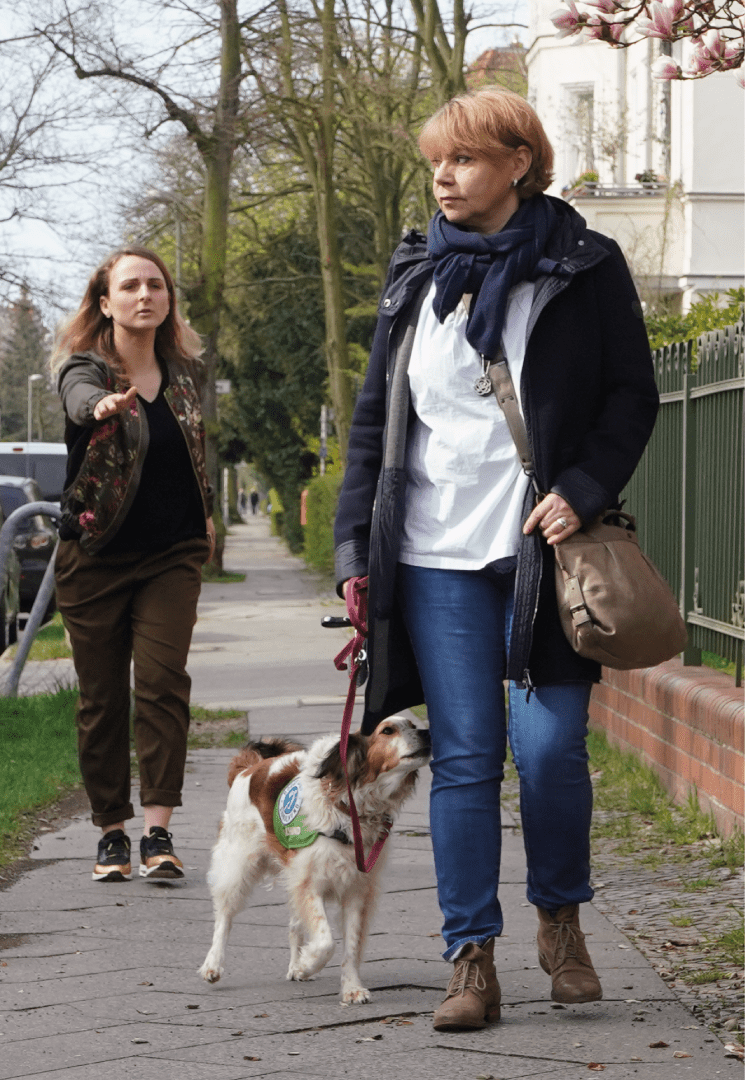 Eine Frau versucht, die Aufmerksamkeit einer anderen Frau zu erlangen. Deren Assistenzhund stupst sie mit der Nase an, um sie auf die Frau aufmerksam zu machen, die versucht, ihre Aufmerksamkeit zu erlangen.