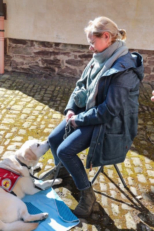 Eine Frau sitzt auf einem kleinen Klapphocker und der Assistenzhund liegt davor