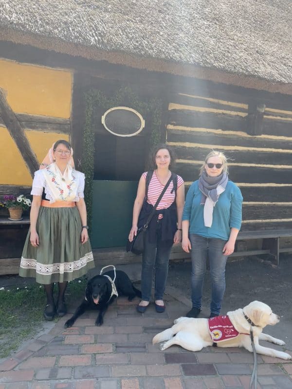 Zwei Assistenzhund-Mensch-Teams stehen vor einem alten Haus im Freiluftmuseum mit einer Frau in sorbischer Tracht