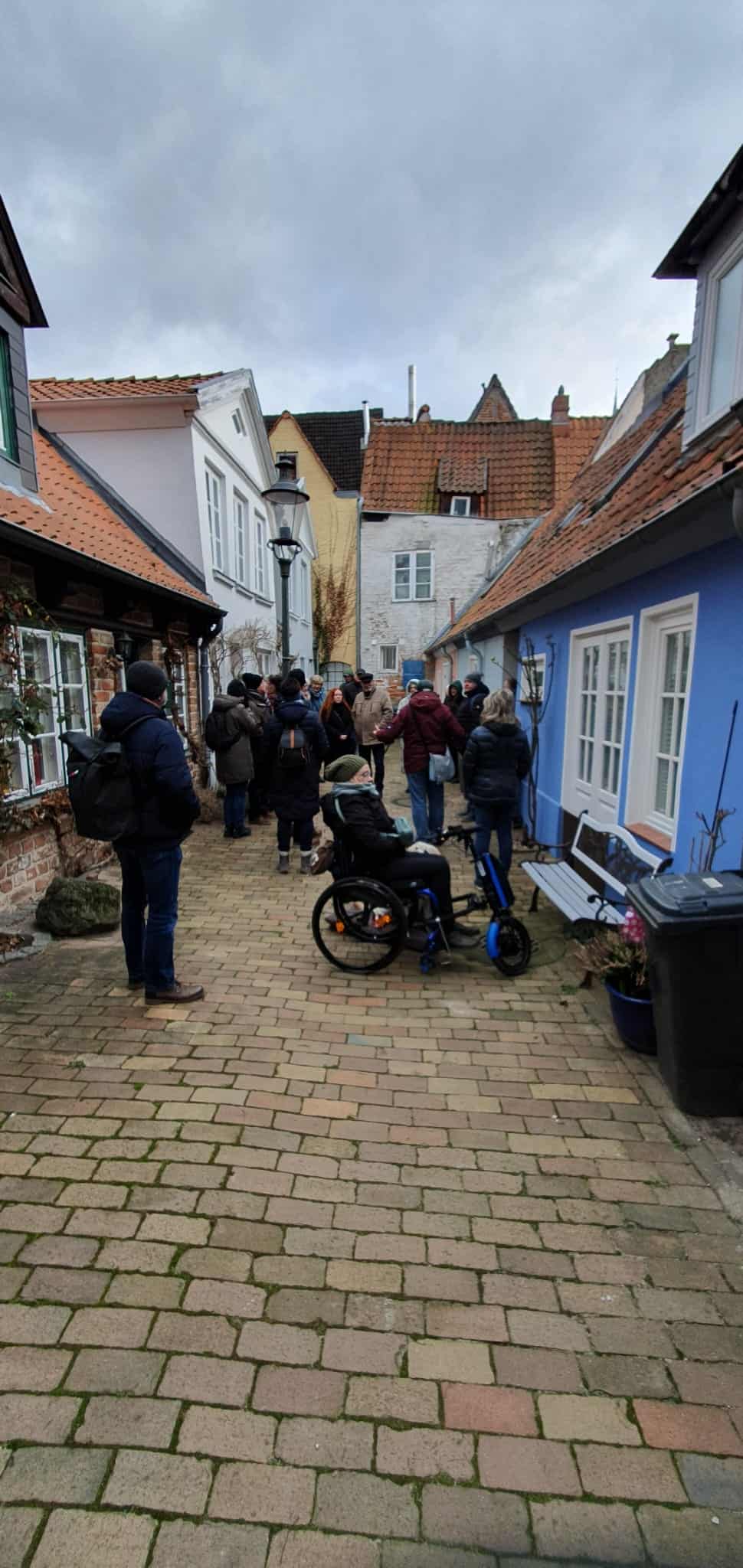 In einem engen Hof steht eine Gruppe von menschen, darunter eine Frau im Rolli mit Assistenzhund