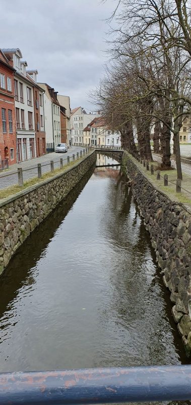 Im Mittelpunkt steht der künstliche Wasserlauf in Wismar, daneben laufen Wege, an die sich historische Häuser anschließen