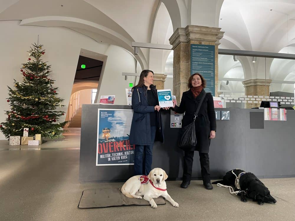 Ines Schnee stands in the foyer of the military history museum with an oversized "Assistance dog welcome" sticker and Dr. Hannah Reuter with the assistance dogs Mascha and Daika, beaming at the camera.