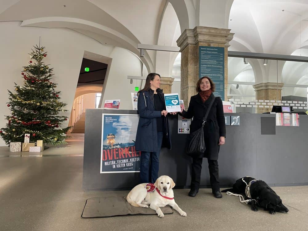 Im Foyer des militärhistorischen Museums stehen Ines Schnee mit einem übergroßen „Assistenzhund willkommen“ Aufkleber und Dr. Hannah Reuter mit den Assistenzhunden Mascha und Daika und strahlen in die Kamera.