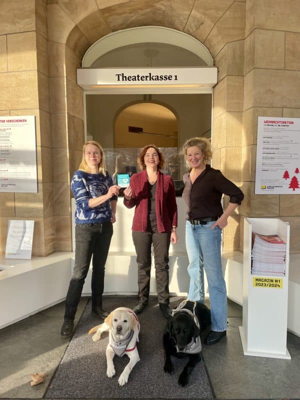 An der Theaterkasse des Staatsschauspiel Dresden halten Franziska Blech (Leitung Presse- und Öffentlichkeitsarbeit) und Janny Fuchs (Theaterpädagogik) den Aufkleber „Assistenzhund willkommen) hoch. Im Vordergrund liegen Daika und Mascha.