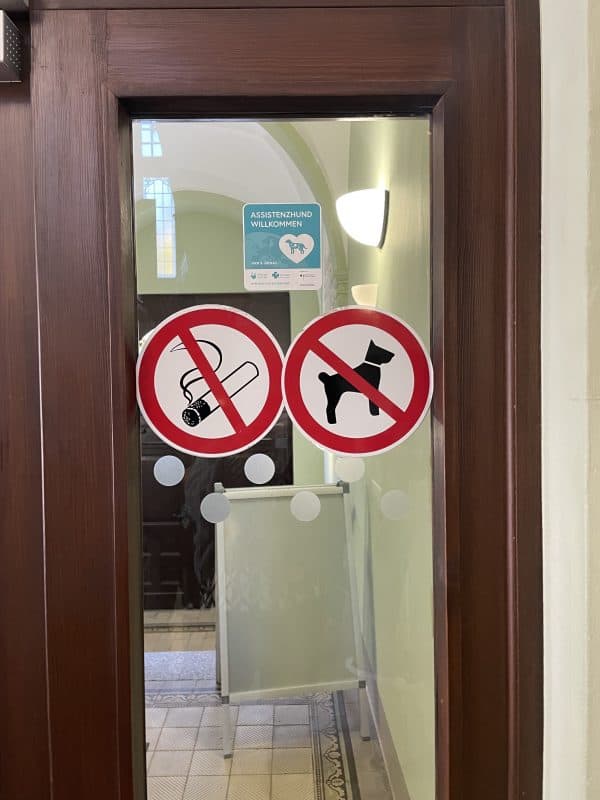 An der Glastür des Bürgeramtes Leuben sieht man die Piktogramme für Rauchverbot und Hundeverbot. Über beiden wurde der Aufkleber „Assistenzhund willkommen“ platziert.