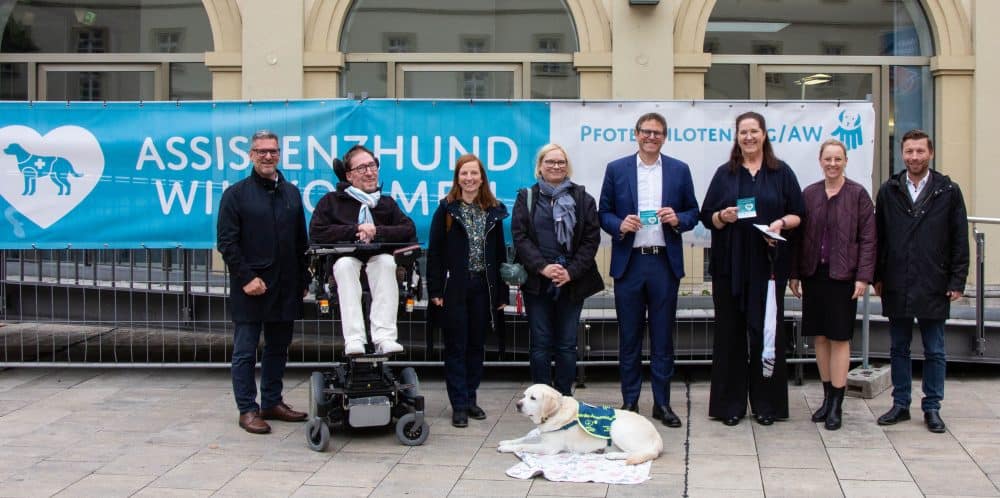 Eine Gruppe von Menschen steht in einer Reihe vor einem großen, weiß/blauen Banner der Pfotenpiloten zum Thema Assistenzhund willkommen.
Ein heller Labrador liegt mit Kenndecke vor der Gruppe.