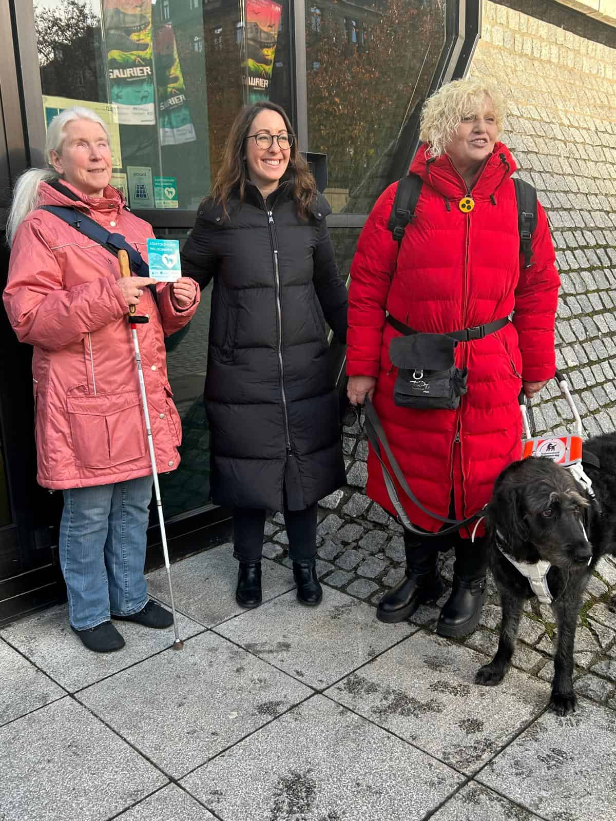 Drei Damen stehen freundlich lächelnd vor dem Eingang des Museums.  Die Frau links hält ihren Blindenstock und den Aufkleber „Assistenzhund willkommen“ in den Händen. In der Mitte steht eine Frau mit dunkler Kleidung, braunen Haaren und Brille. Rechts daneben steht eine blonde Frau mit rotem Mantel und ihrem Blindenführhund am Geschirr.